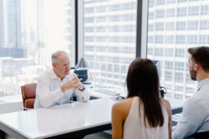 A man in a collared shirt sits at a desk talking to a young couple.